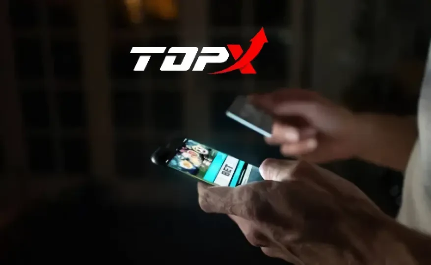 Mãos segurando um celular aceso em ambiente escuro, com a marca TopX